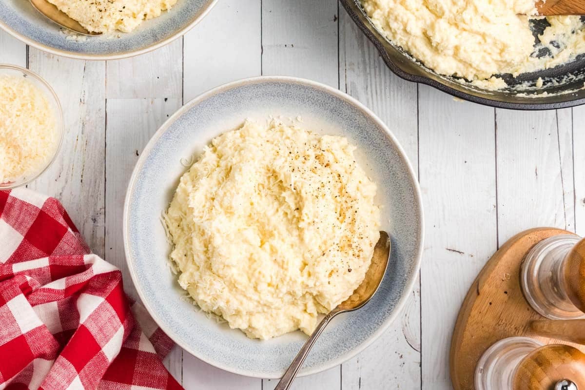 A bowl of creamy baked polenta with a spoon, topped with ground pepper, on a white wooden table next to a red checkered napkin and other bowls of mashed potatoes.