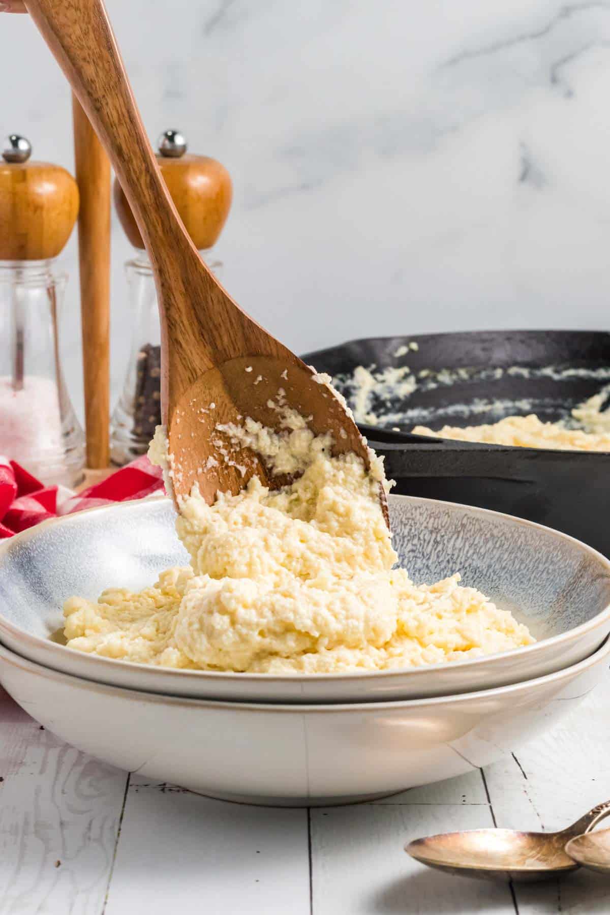 A wooden spoon scoops baked polenta from a skillet into a white bowl, with salt and pepper shakers and a spoon visible in the background.