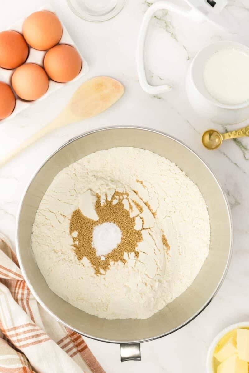 A mixing bowl with flour, yeast, and baking soda, surrounded by eggs, milk, butter, a wooden spoon, a kitchen towel, and measuring spoons on a white countertop.