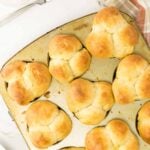 A muffin tin filled with nine freshly baked cloverleaf dinner rolls sits on a white surface next to a knife and a plaid kitchen towel.