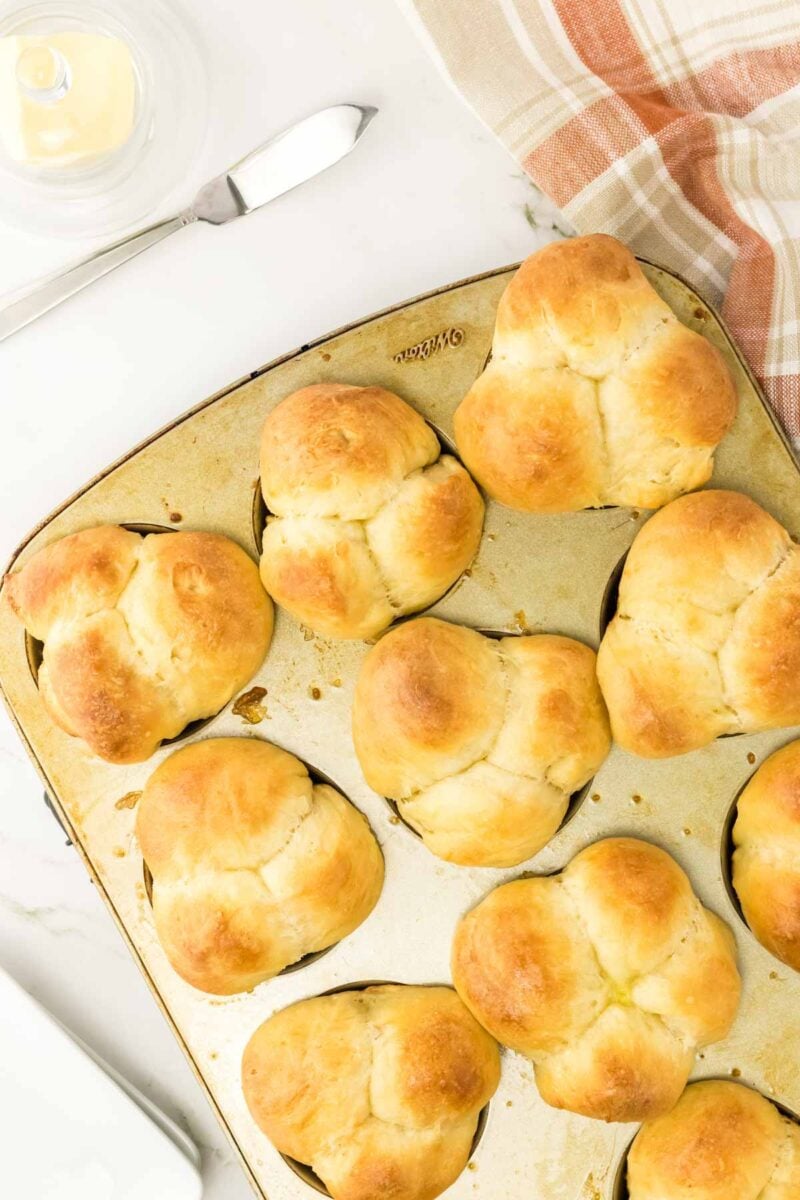 A muffin tin filled with nine freshly baked cloverleaf dinner rolls sits on a white surface next to a knife and a plaid kitchen towel.