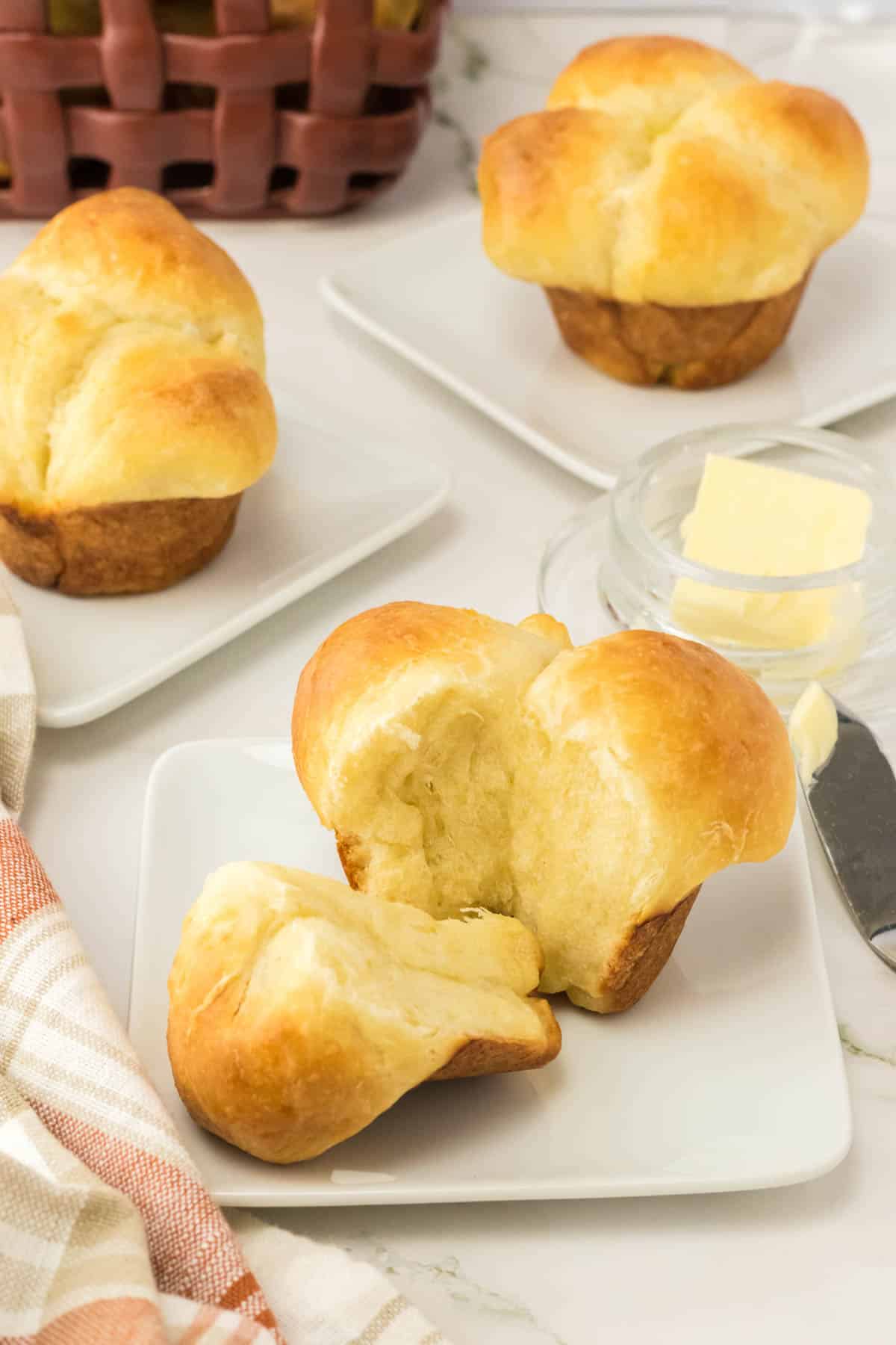 Three golden brown cloverleaf rolls sit on square white plates; one is split open. A small glass dish of butter and a butter knife are nearby on the table.