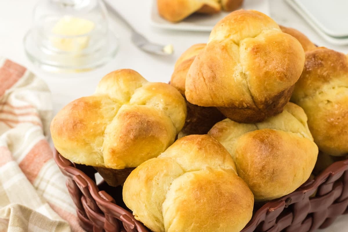 A basket filled with golden brown cloverleaf rolls, with a striped cloth underneath and butter in the background.