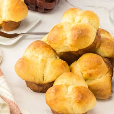 A pile of golden brown cloverleaf dinner rolls sits on a marble surface, with a plate, butter knife, and napkin nearby.