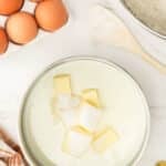 A saucepan with milk, butter, and sugar next to a bowl of flour, a wooden spoon, eggs in a carton, and a striped towel on a marble countertop.