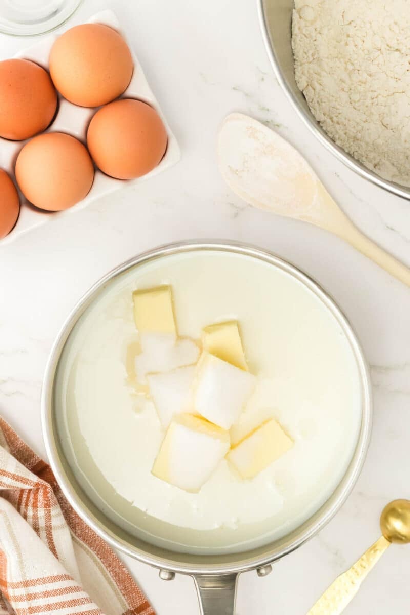 A saucepan with milk, butter, and sugar next to a bowl of flour, a wooden spoon, eggs in a carton, and a striped towel on a marble countertop.