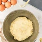 A metal bowl with risen dough sits on a counter next to brown eggs, a dough scraper, a spoon, and a striped kitchen towel.