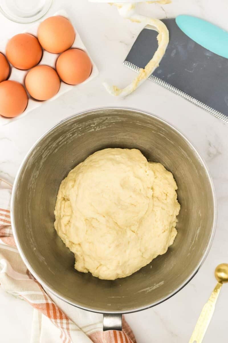 A metal bowl with risen dough sits on a counter next to brown eggs, a dough scraper, a spoon, and a striped kitchen towel.