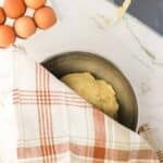 A metal bowl with dough covered by a plaid kitchen towel sits on a marble surface next to a carton of eggs and a dough scraper with residue.
