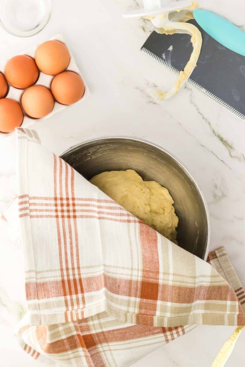A metal bowl with dough covered by a plaid kitchen towel sits on a marble surface next to a carton of eggs and a dough scraper with residue.