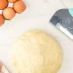 Ball of dough on a floured surface with a dough scraper, a carton of eggs, a jar of flour, and a striped towel nearby.