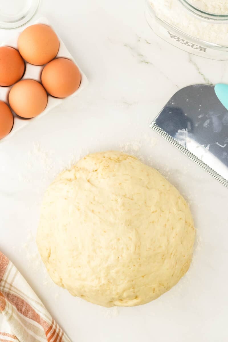 Ball of dough on a floured surface with a dough scraper, a carton of eggs, a jar of flour, and a striped towel nearby.
