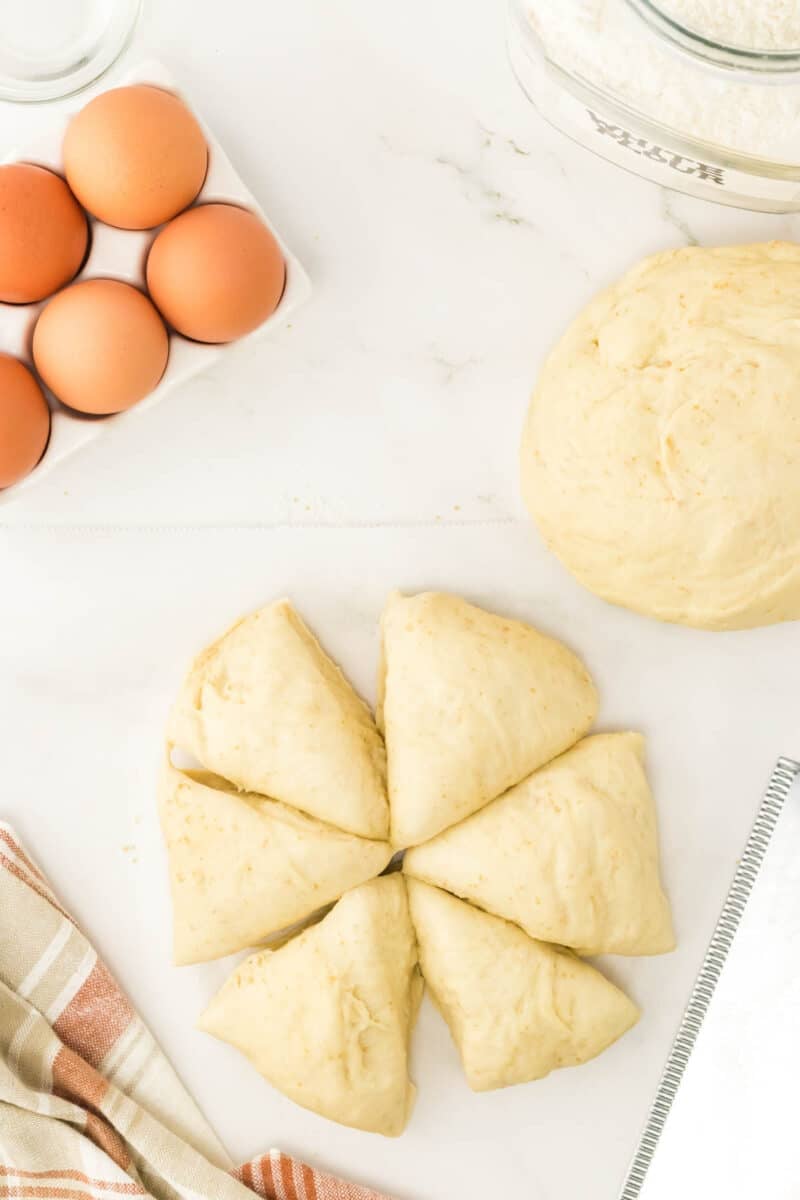 Sliced dough pieces arranged in a circle, a ball of dough, a carton of eggs, a jar of flour, and a striped towel on a white surface.