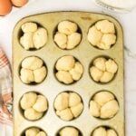 A metal muffin pan filled with unbaked cloverleaf rolls sits on a counter next to a jar of flour, six eggs, and a striped kitchen towel.