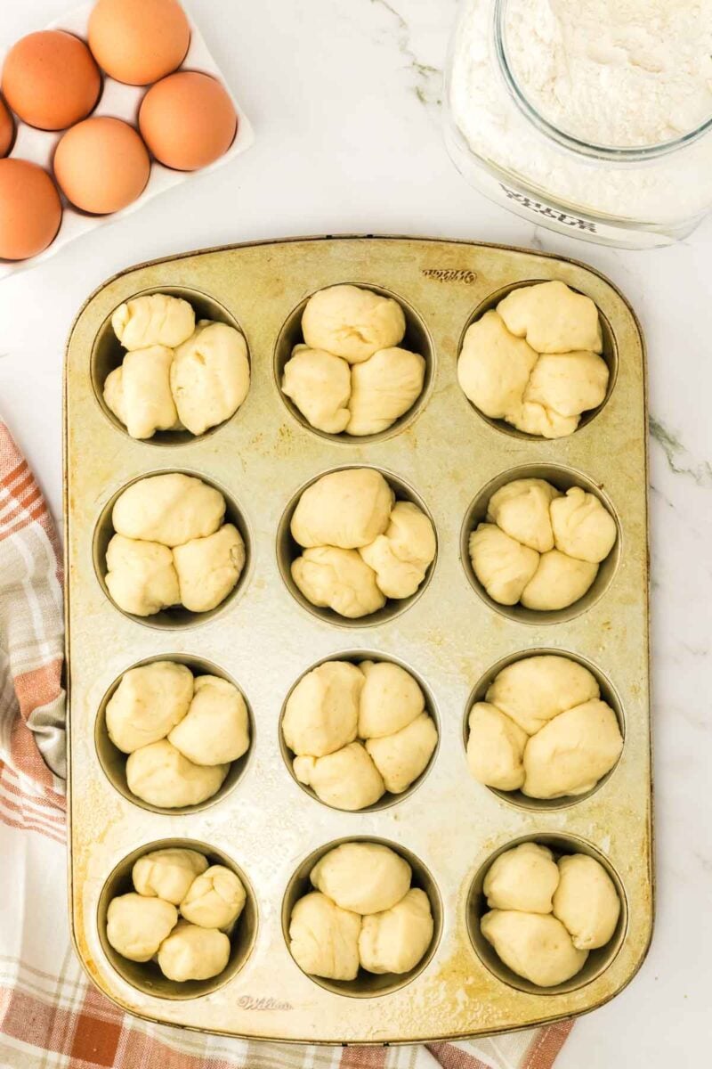 A metal muffin pan filled with unbaked cloverleaf rolls sits on a counter next to a jar of flour, six eggs, and a striped kitchen towel.