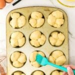A hand brushes dough with egg wash in a muffin tin, with whole eggs, a bowl of beaten egg, and a fork on a marble countertop nearby.