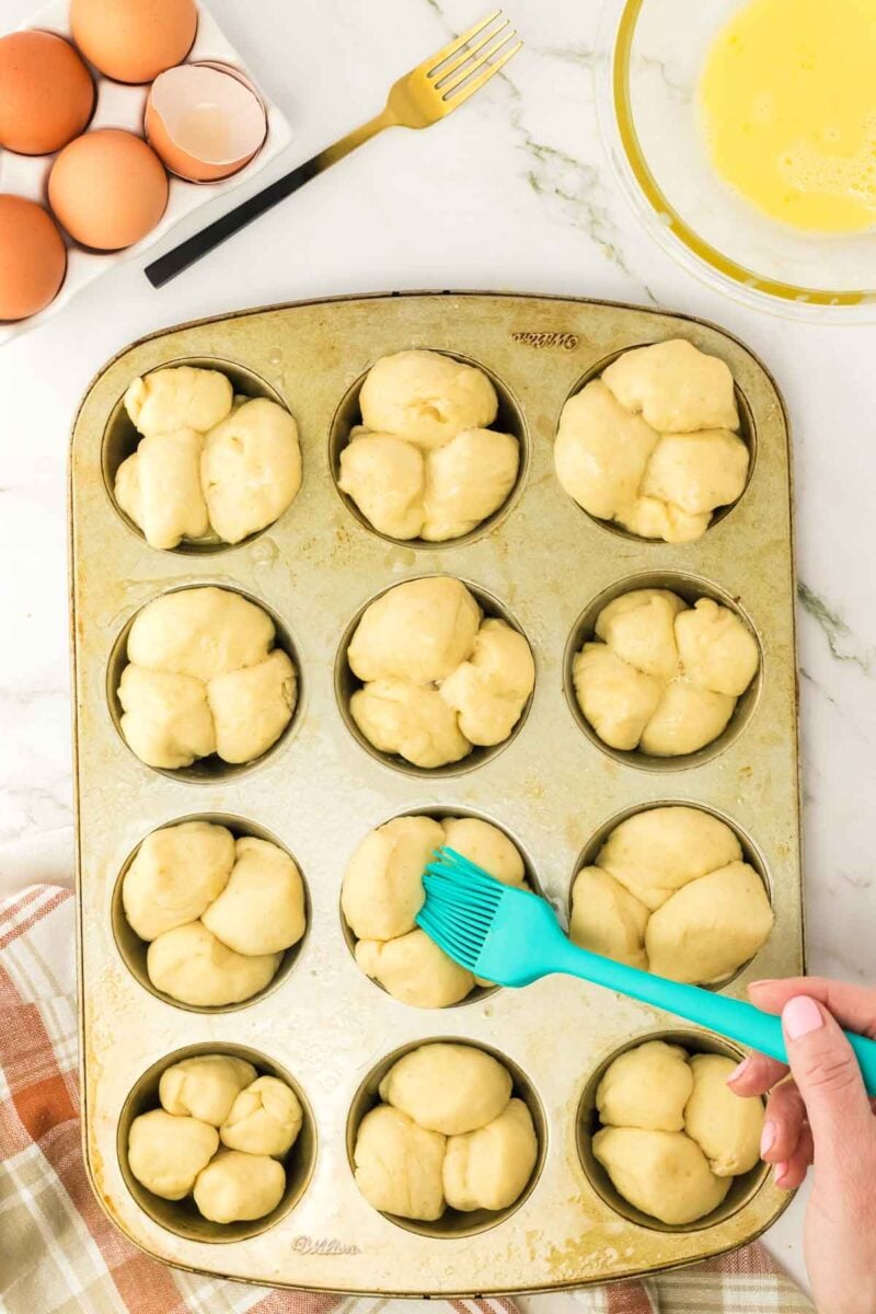 A hand brushes dough with egg wash in a muffin tin, with whole eggs, a bowl of beaten egg, and a fork on a marble countertop nearby.
