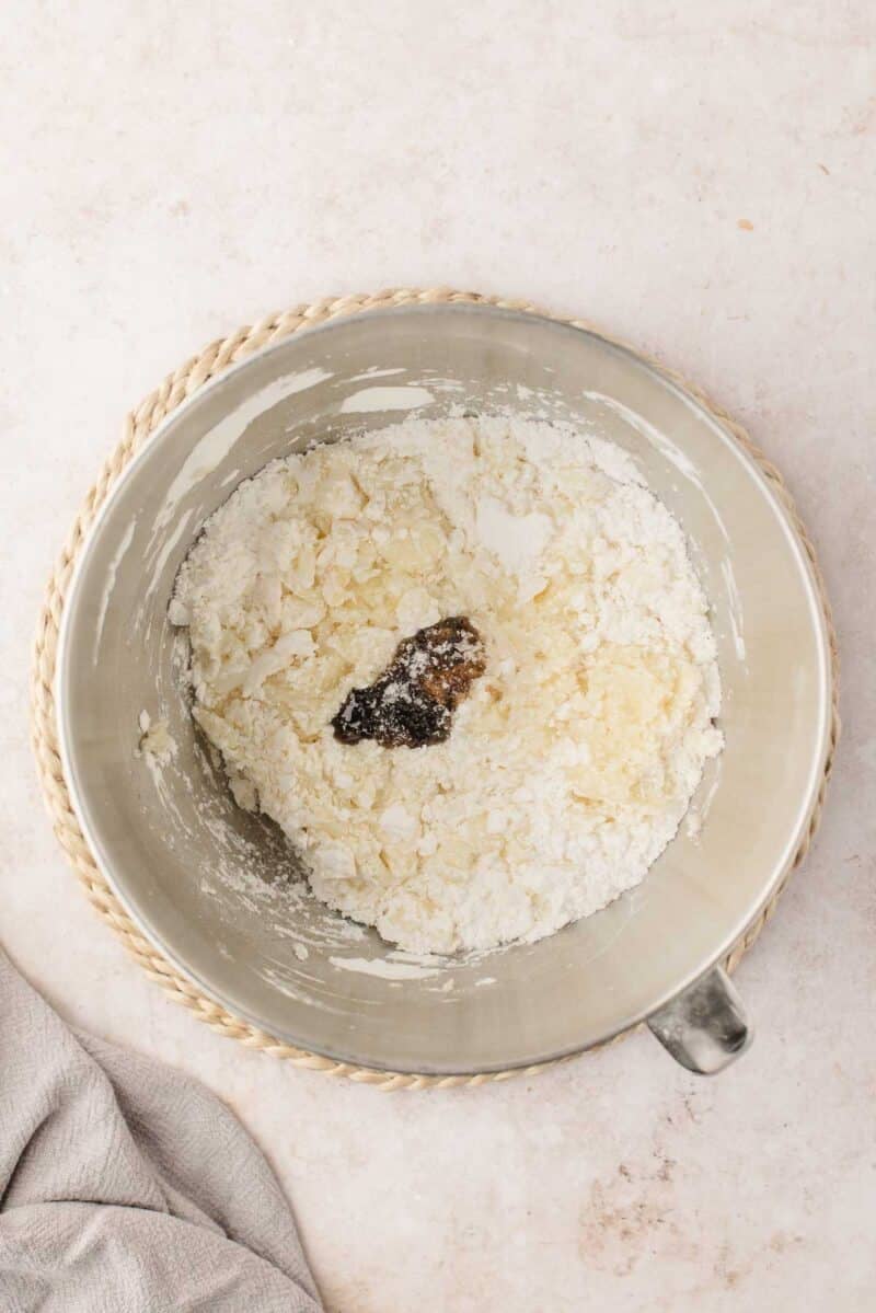 A mixing bowl containing flour, sugar, butter, and vanilla extract, partially mixed, on a light countertop with a cloth nearby.