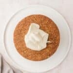 A round, unfrosted cake sits on a white plate with a dollop of white frosting on top, next to a light-colored textured cloth.