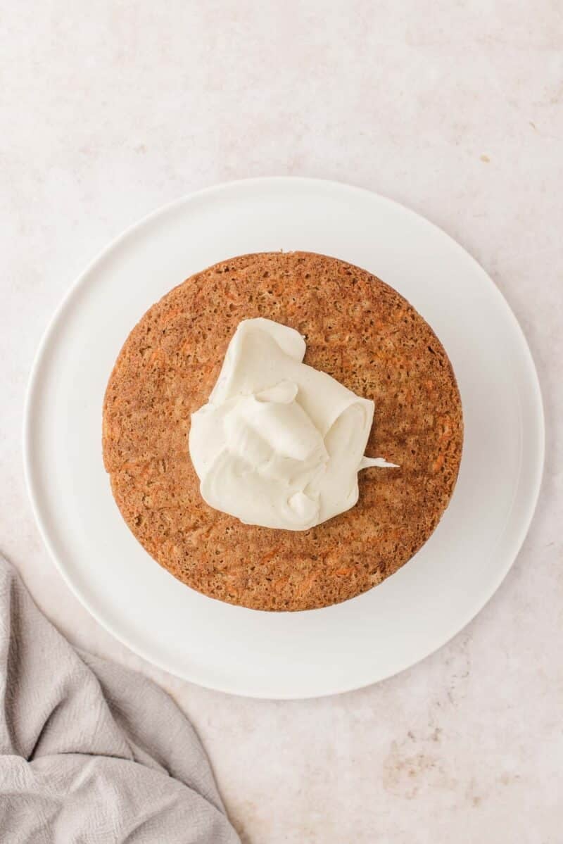 A round, unfrosted cake sits on a white plate with a dollop of white frosting on top, next to a light-colored textured cloth.