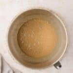 A metal mixing bowl filled with a light brown, frothy liquid sits on a woven mat next to a beige cloth on a light countertop.