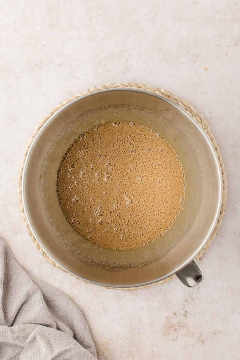A metal mixing bowl filled with a light brown, frothy liquid sits on a woven mat next to a beige cloth on a light countertop.