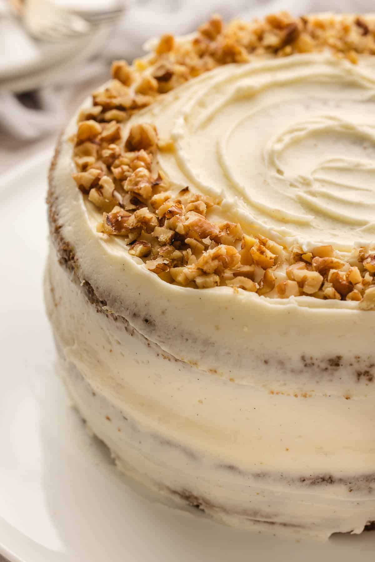 A frosted carrot layer cake topped with chopped walnuts sits on a white plate.
