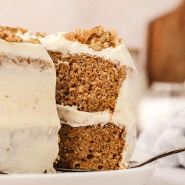 A close-up of a frosted carrot layer cake with chopped nuts on top, showing a slice being lifted with a cake server.