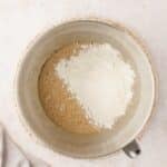 A metal mixing bowl containing a brown liquid mixture and a pile of white flour on top, placed on a woven mat.