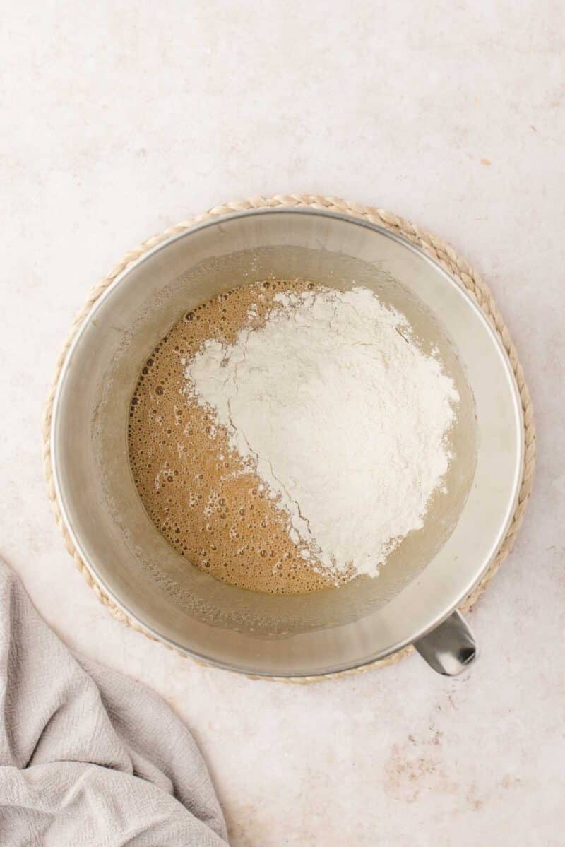 A metal mixing bowl containing a brown liquid mixture and a pile of white flour on top, placed on a woven mat.