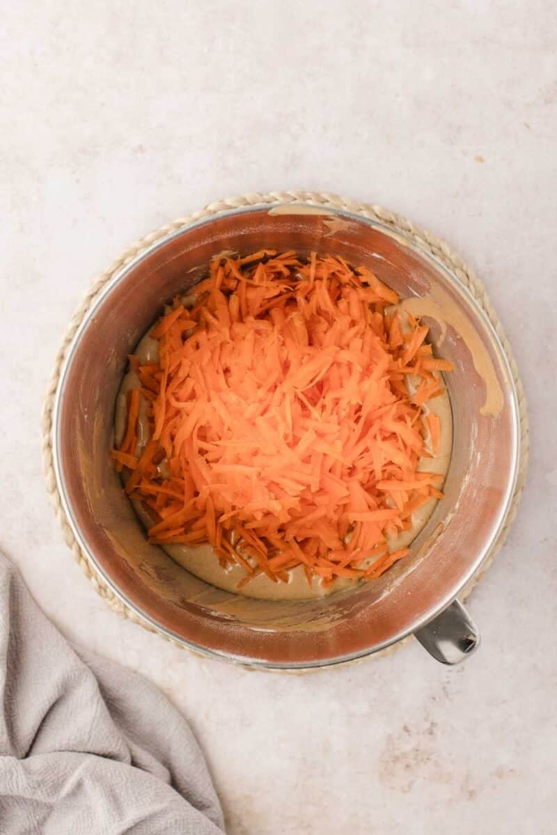 A mixing bowl with shredded carrots on top of a light-colored batter, placed on a woven mat, with a gray cloth nearby.