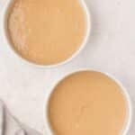 Two round white baking pans filled with light brown cake batter on a light-colored surface, with a beige cloth in the bottom left corner.