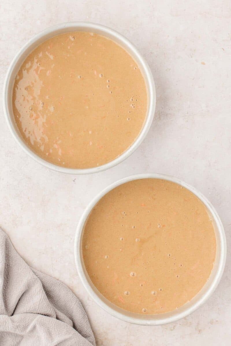 Two round white baking pans filled with light brown cake batter on a light-colored surface, with a beige cloth in the bottom left corner.