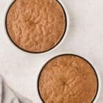 Two round chocolate cakes in white baking pans on a light-colored surface, with a light gray cloth partially visible in the corner.