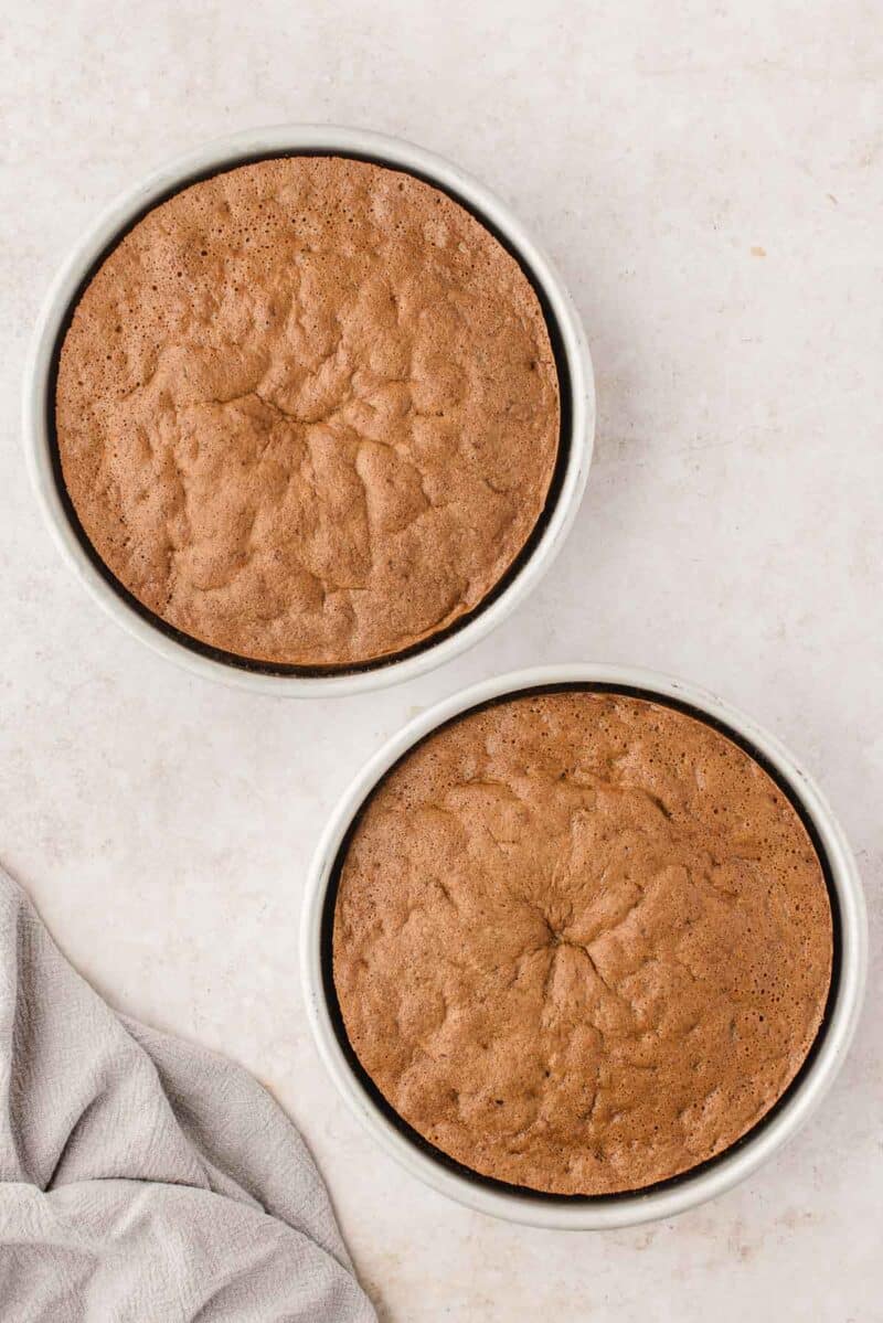 Two round chocolate cakes in white baking pans on a light-colored surface, with a light gray cloth partially visible in the corner.