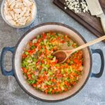 A pot with chopped celery, carrots, and onions being stirred with a wooden spoon, next to a bowl of shredded chicken and a cutting board with minced garlic and a knife.