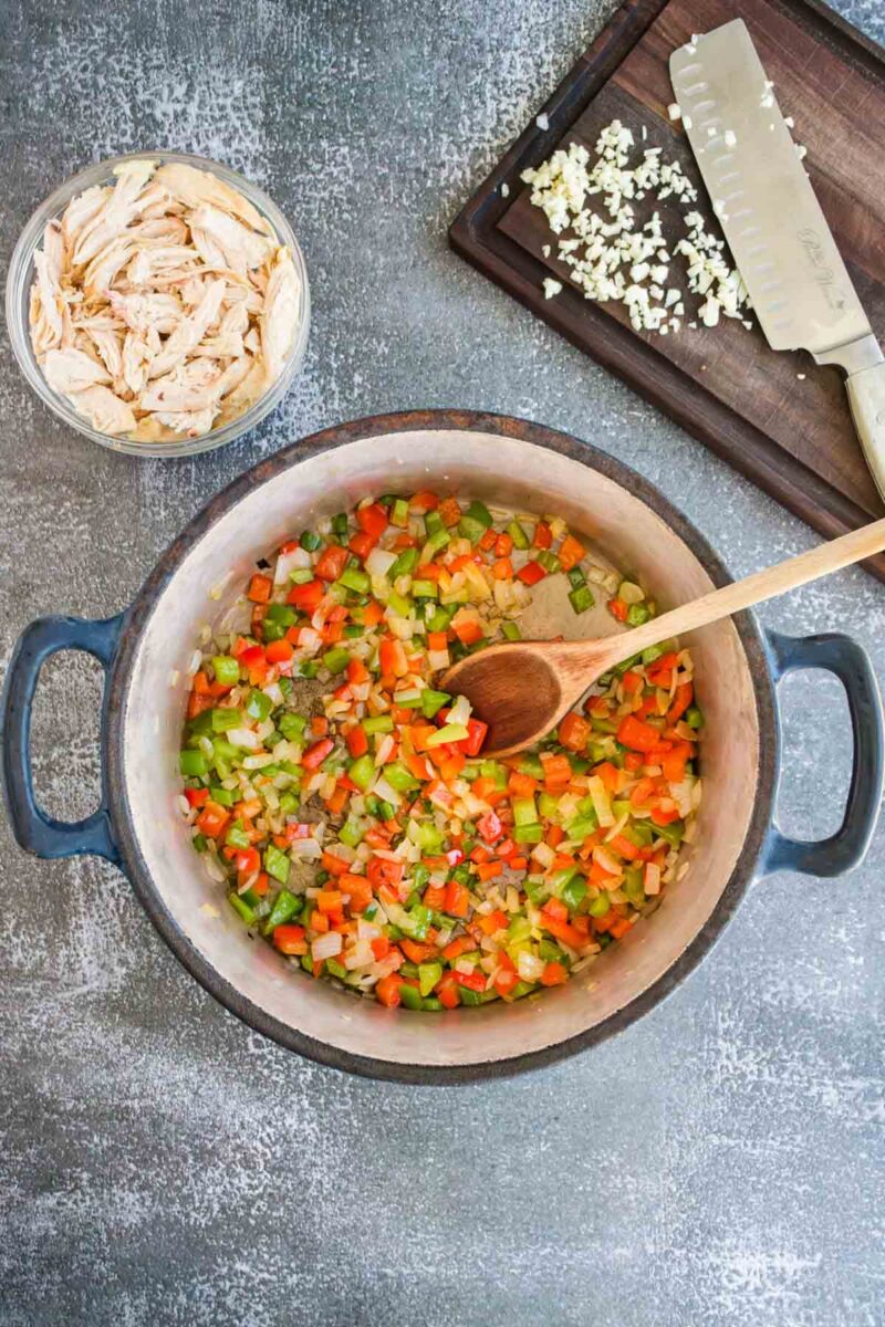 A pot with chopped celery, carrots, and onions being stirred with a wooden spoon, next to a bowl of shredded chicken and a cutting board with minced garlic and a knife.