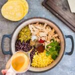 A pot containing shredded chicken, black beans, corn, green chiles, cilantro, and spices is being filled with broth. Yellow corn tortillas and a cutting board are next to the pot.