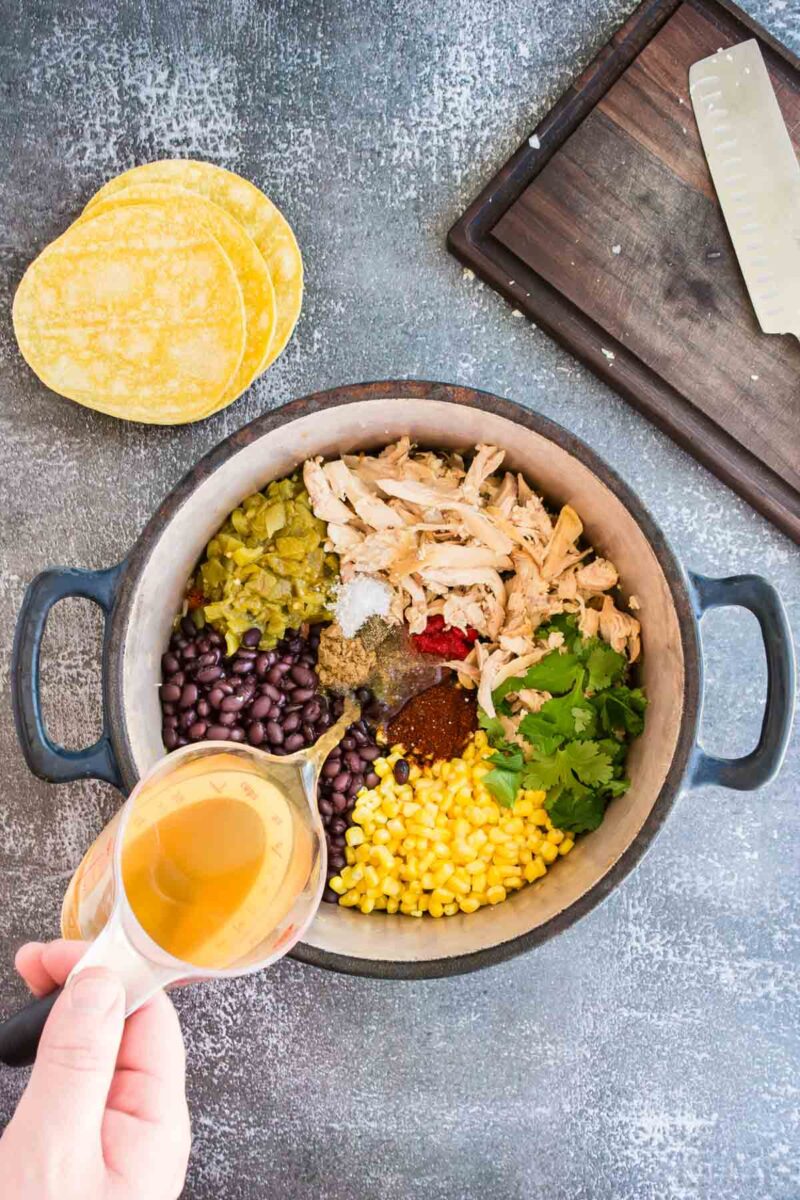 A pot containing shredded chicken, black beans, corn, green chiles, cilantro, and spices is being filled with broth. Yellow corn tortillas and a cutting board are next to the pot.