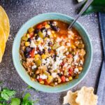 A bowl of vegetable soup topped with crumbled cheese, surrounded by tortilla chips, lime wedges, cilantro, and corn tortillas on a gray surface.