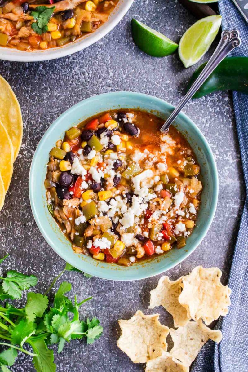 A bowl of vegetable soup topped with crumbled cheese, surrounded by tortilla chips, lime wedges, cilantro, and corn tortillas on a gray surface.