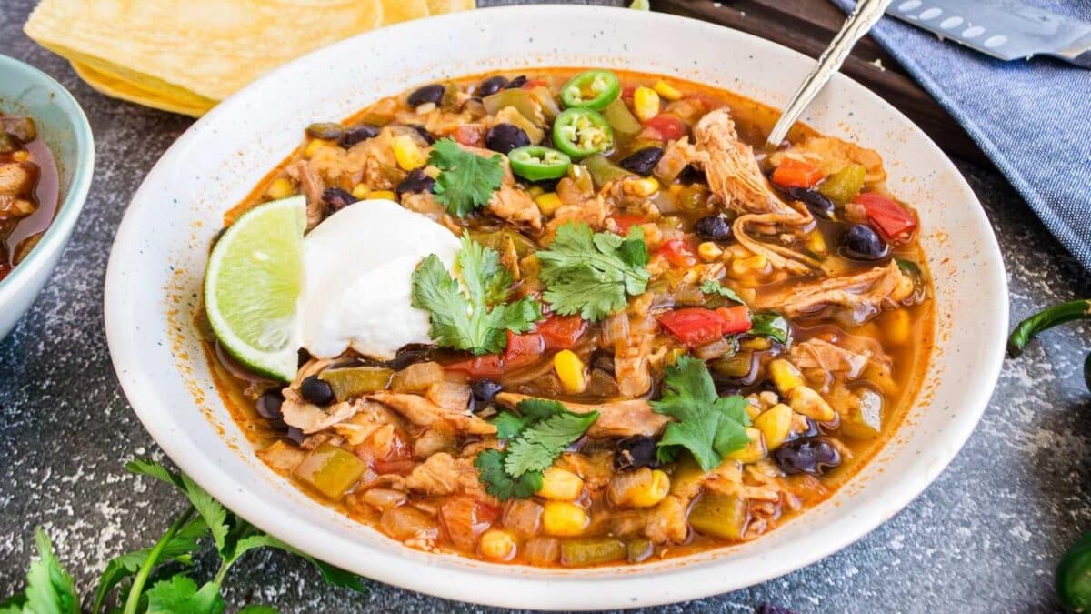 A bowl of chicken chili with black beans, corn, diced tomatoes, and peppers, topped with sour cream, cilantro, lime, and sliced jalapeños.
