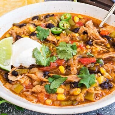 A bowl of chicken chili with black beans, corn, diced tomatoes, and peppers, topped with sour cream, cilantro, lime, and sliced jalapeรฑos.