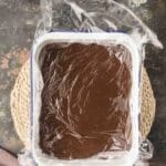 A square dish filled with chocolate mixture covered by plastic wrap, placed on a woven mat on a dark, textured surface.