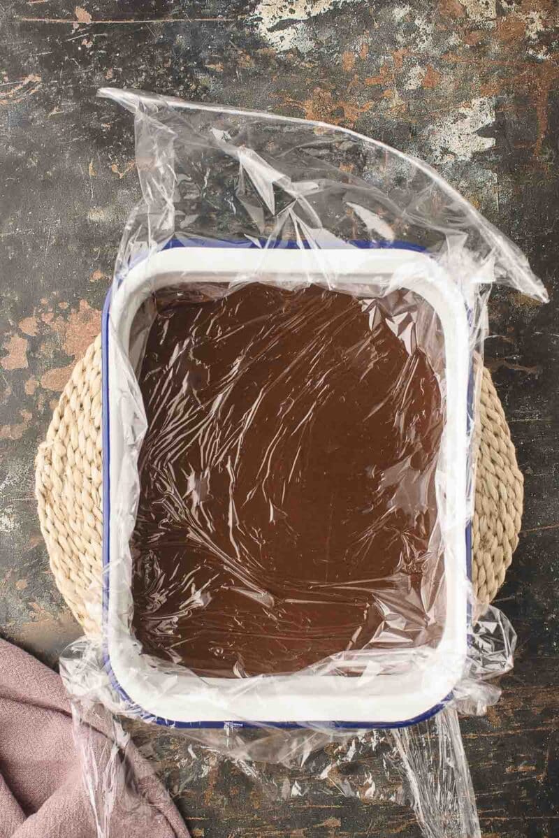 A square dish filled with chocolate mixture covered by plastic wrap, placed on a woven mat on a dark, textured surface.