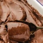 A metal spoon scoops a portion of smooth, glossy chocolate cremeux from a pan covered with thick, swirled chocolate frosting.