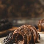 A chocolate cremeux with a bite taken out, garnished with chocolate shavings and served with fresh blackberries on a ceramic plate.