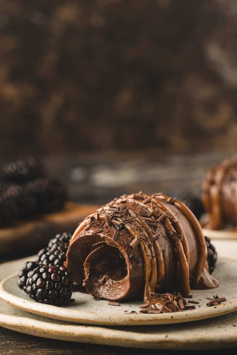 A chocolate cremeux with a bite taken out, garnished with chocolate shavings and served with fresh blackberries on a ceramic plate.