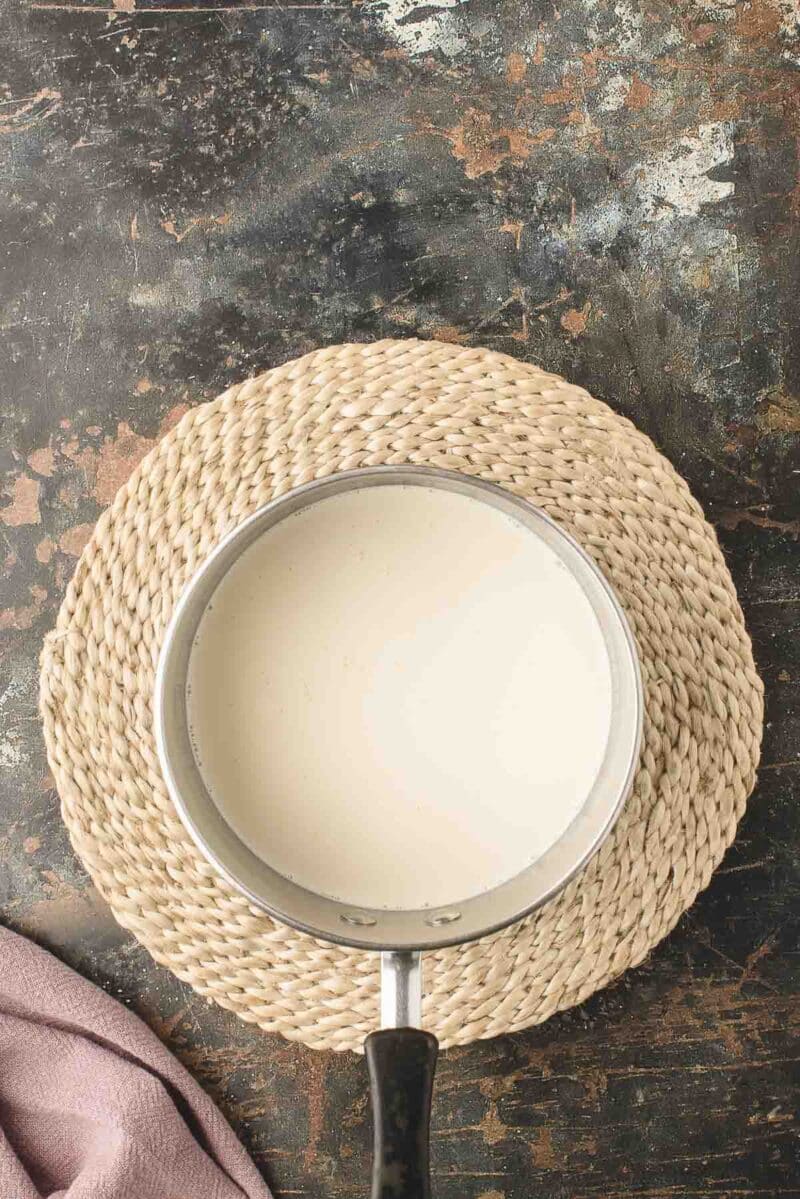 A saucepan filled with milk sits on a round woven placemat atop a dark, textured surface.