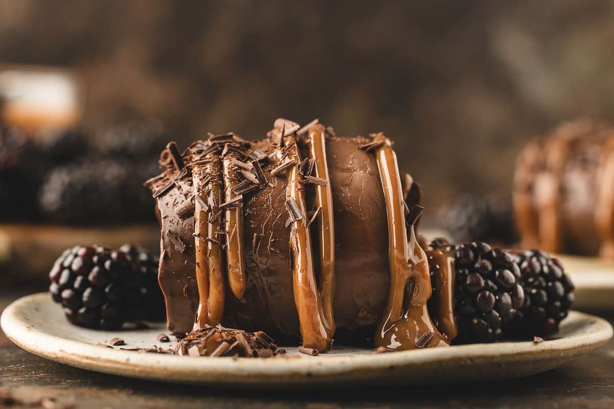 A chocolate cremeux topped with caramel sauce and chocolate shavings, served on a plate with blackberries.